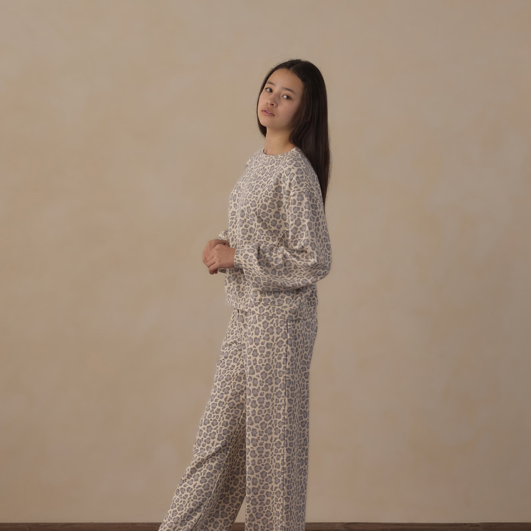 A woman stands barefoot in a studio setting wearing a matching two-piece pajama set with a beige and brown abstract pattern. The set includes a long-sleeve top and wide-leg pants, set against a neutral beige wall and dark wood floor.