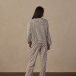 A woman stands with her back to the camera wearing a matching two-piece pajama set with a beige and brown animal print pattern, set against a plain beige wall and dark wood floor.