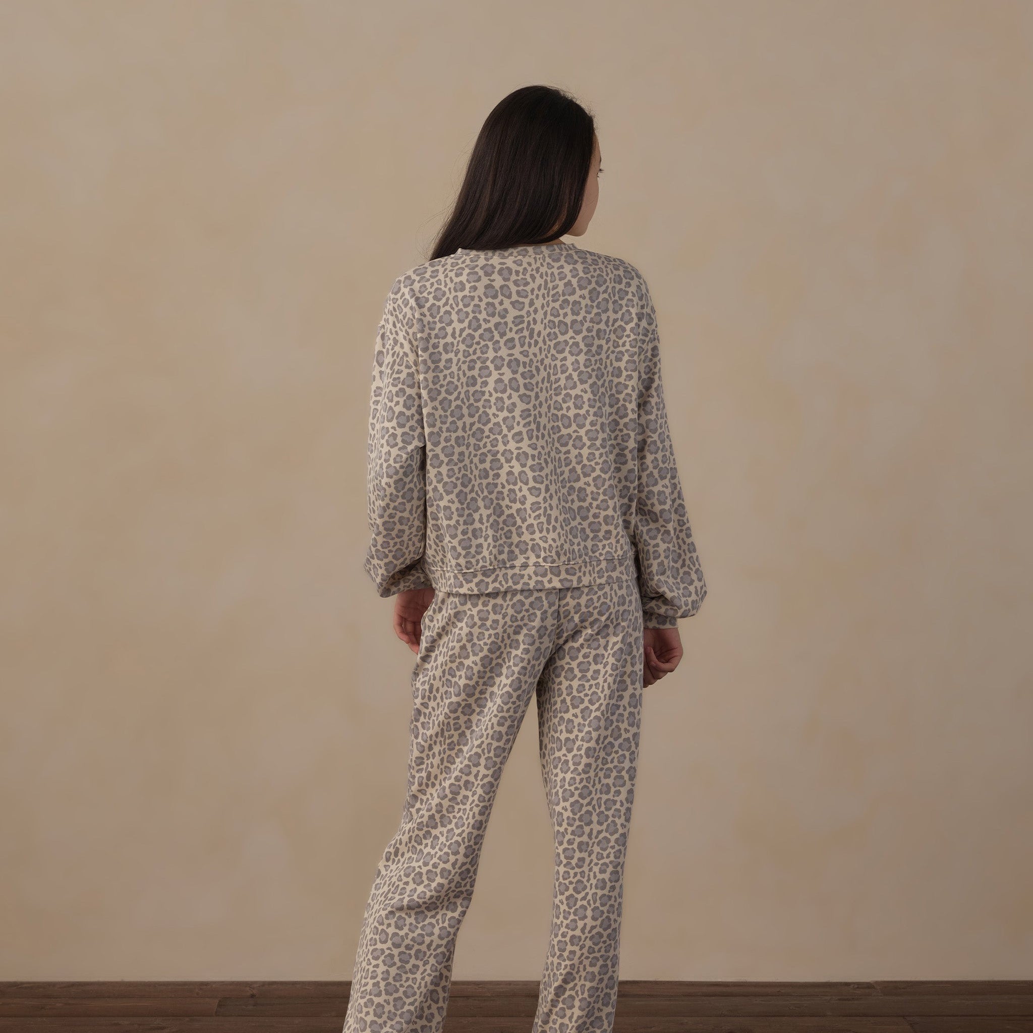 A woman stands with her back to the camera wearing a matching two-piece pajama set with a beige and brown animal print pattern, set against a plain beige wall and dark wood floor.