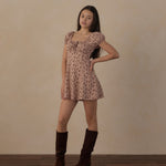 A woman modeling a short puff-sleeve dress with a small floral print in dusty pink and brown, paired with dark brown knee-high boots, standing against a textured beige wall on a wooden floor.
