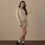A young woman models a cream-colored textured knit cardigan and matching crochet mini skirt set, styled with white socks and black loafers against a neutral beige wall and wooden floor.