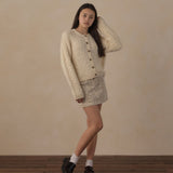 A young woman models a cream-colored textured knit cardigan and matching crochet mini skirt set, styled with white socks and black loafers against a neutral beige wall and wooden floor.