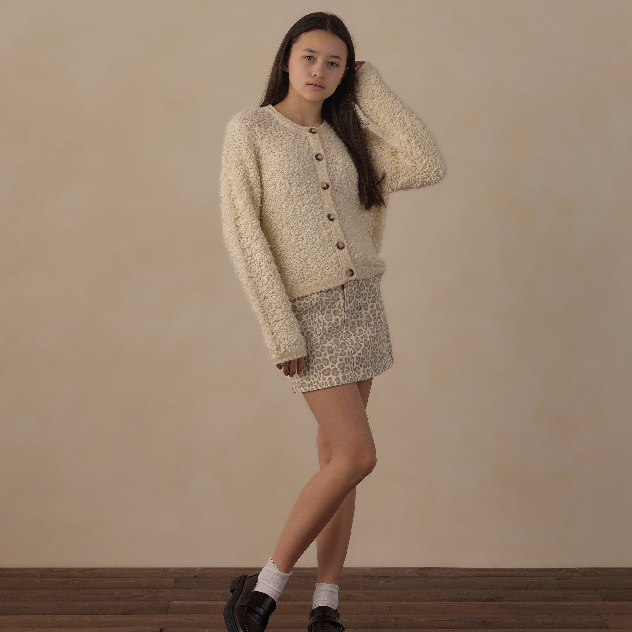 A young woman models a cream-colored textured knit cardigan and matching crochet mini skirt set, styled with white socks and black loafers against a neutral beige wall and wooden floor.