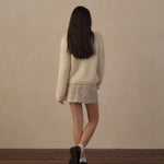 A woman is shown from behind wearing a cream-colored fuzzy long-sleeve sweater and a beige-and-black patterned mini skirt, standing on a dark wood floor against a textured beige wall.