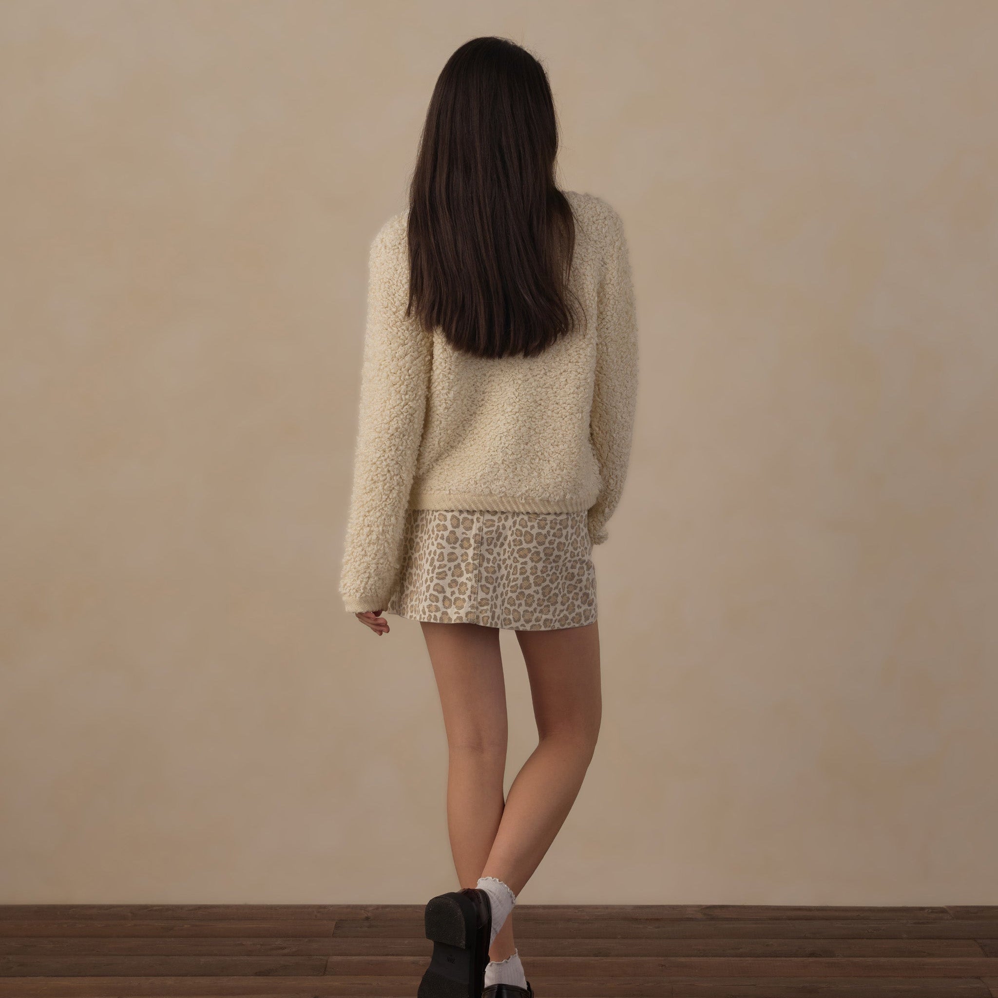 A woman is shown from behind wearing a cream-colored fuzzy long-sleeve sweater and a beige-and-black patterned mini skirt, standing on a dark wood floor against a textured beige wall.