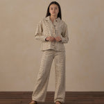 A woman stands in a studio wearing a matching two-piece pajama set with a beige and brown animal print. The set includes a long-sleeve button-up shirt and wide-leg pants, paired with tan slip-on shoes against a textured beige backdrop and dark wood flooring.