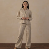 A woman stands in a studio wearing a matching two-piece pajama set with a beige and brown animal print. The set includes a long-sleeve button-up shirt and wide-leg pants, paired with tan slip-on shoes against a textured beige backdrop and dark wood flooring.