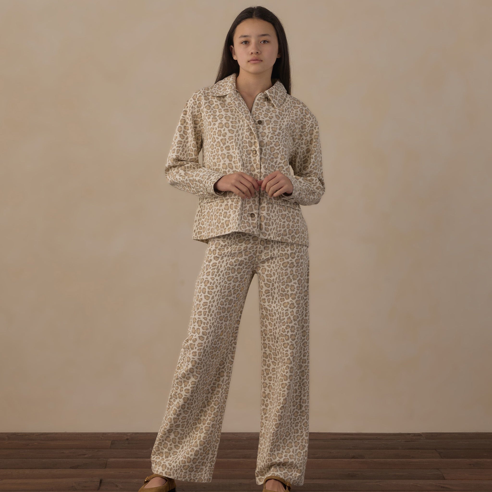 A woman stands in a studio wearing a matching two-piece pajama set with a beige and brown animal print. The set includes a long-sleeve button-up shirt and wide-leg pants, paired with tan slip-on shoes against a textured beige backdrop and dark wood flooring.