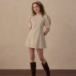 A woman stands in a studio wearing a short puff-sleeve mini dress with an allover small floral print in beige and brown, paired with dark brown knee-high boots and a matching headband. The background is a warm beige textured wall with a dark wood floor.