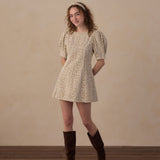 A woman stands in a studio wearing a short puff-sleeve mini dress with an allover small floral print in beige and brown, paired with dark brown knee-high boots and a matching headband. The background is a warm beige textured wall with a dark wood floor.