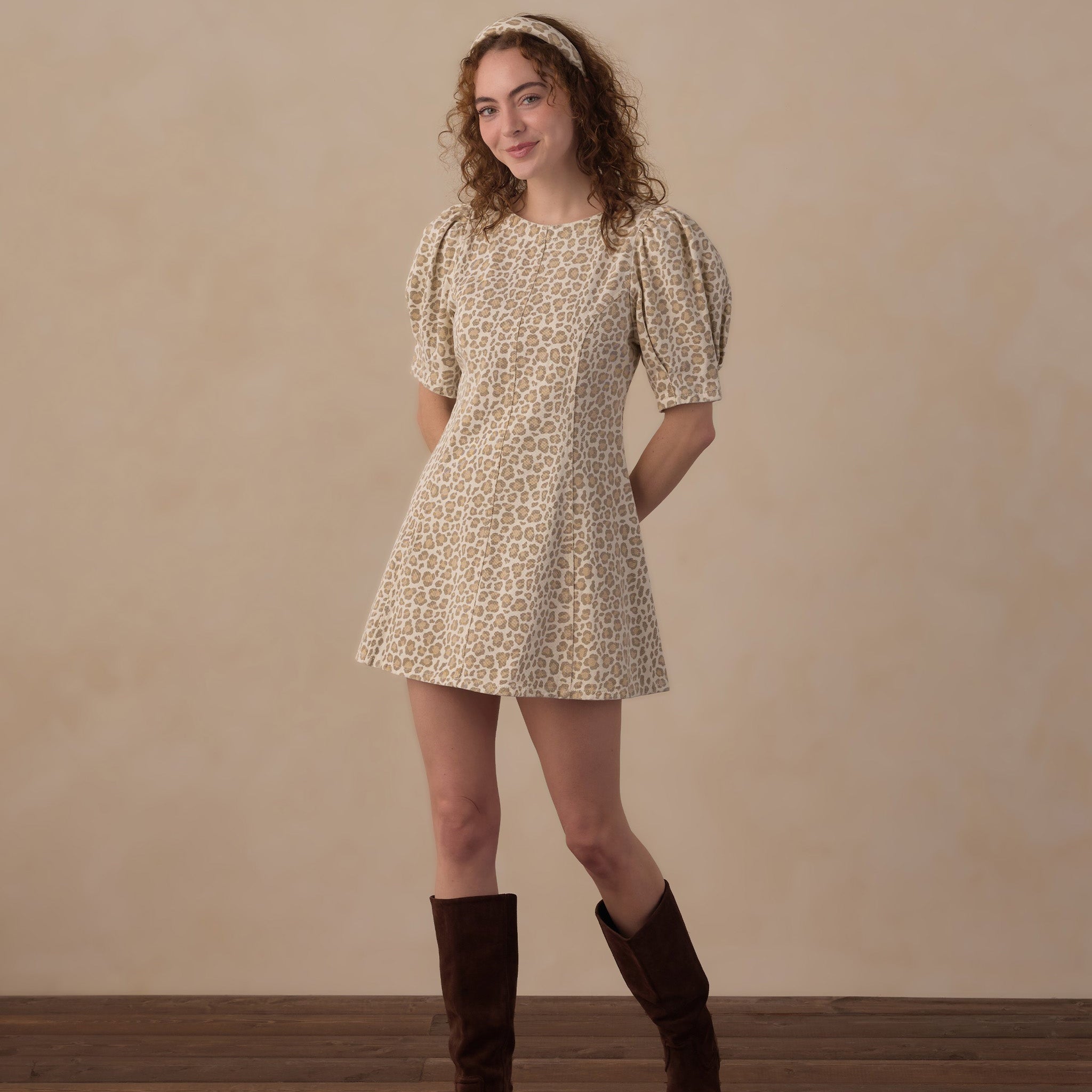 A woman stands in a studio wearing a short puff-sleeve mini dress with an allover small floral print in beige and brown, paired with dark brown knee-high boots and a matching headband. The background is a warm beige textured wall with a dark wood floor.