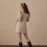 A woman wearing a short-sleeve beige mini dress with a small brown animal print, styled with brown cowboy boots and a matching headband, standing in a studio with a beige wall and wooden floor.