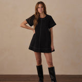 A woman modeling a black short-sleeve mini dress with white contrast stitching, paired with black knee-high boots, standing against a textured beige wall and wooden floor.