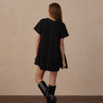 A woman stands with her back to the camera wearing a short-sleeve black dress with white contrast stitching and black knee-high boots, set against a beige wall and dark wood floor.