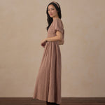 A woman wearing a brown and beige gingham maxi dress with short puff sleeves and a matching headband, standing in profile against a textured beige wall and dark wood floor.
