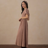 A woman wearing a brown and beige gingham maxi dress with short puff sleeves and a matching headband, standing in profile against a textured beige wall and dark wood floor.
