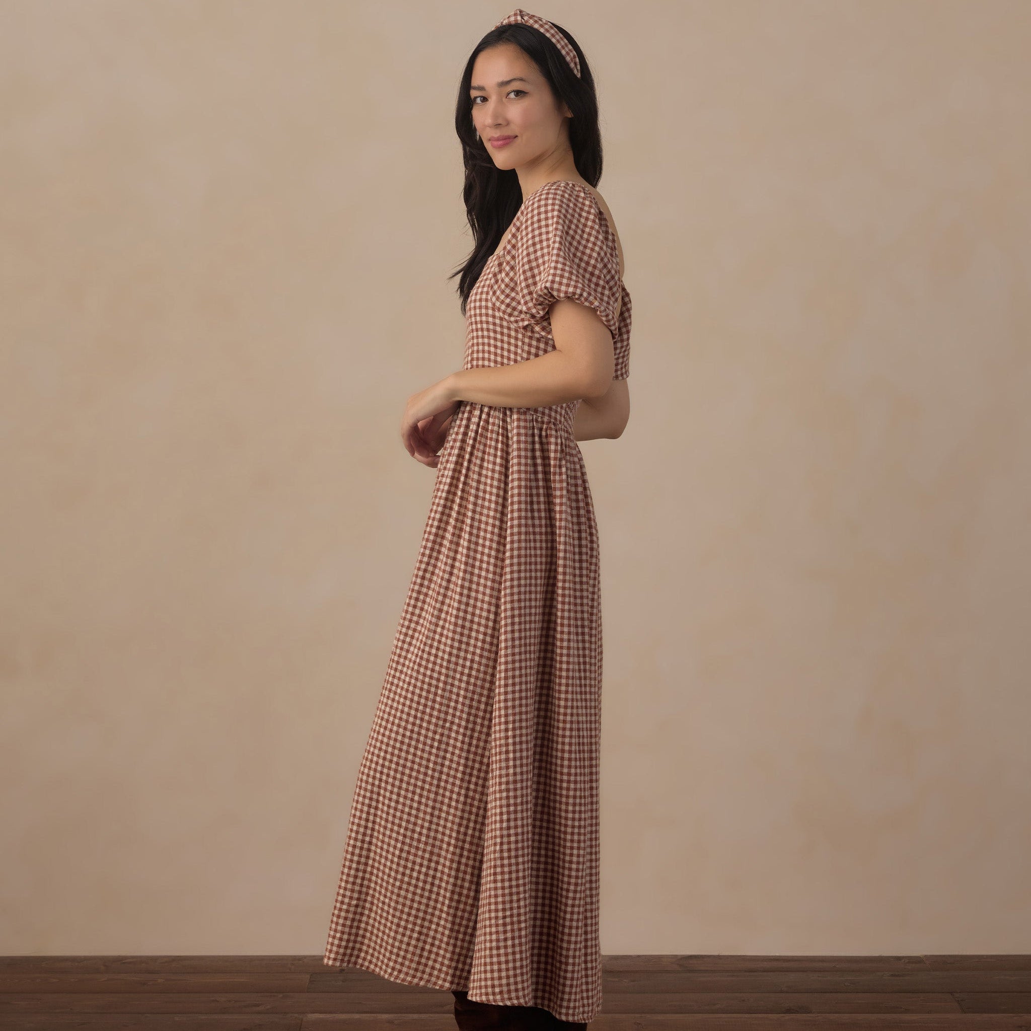 A woman wearing a brown and beige gingham maxi dress with short puff sleeves and a matching headband, standing in profile against a textured beige wall and dark wood floor.