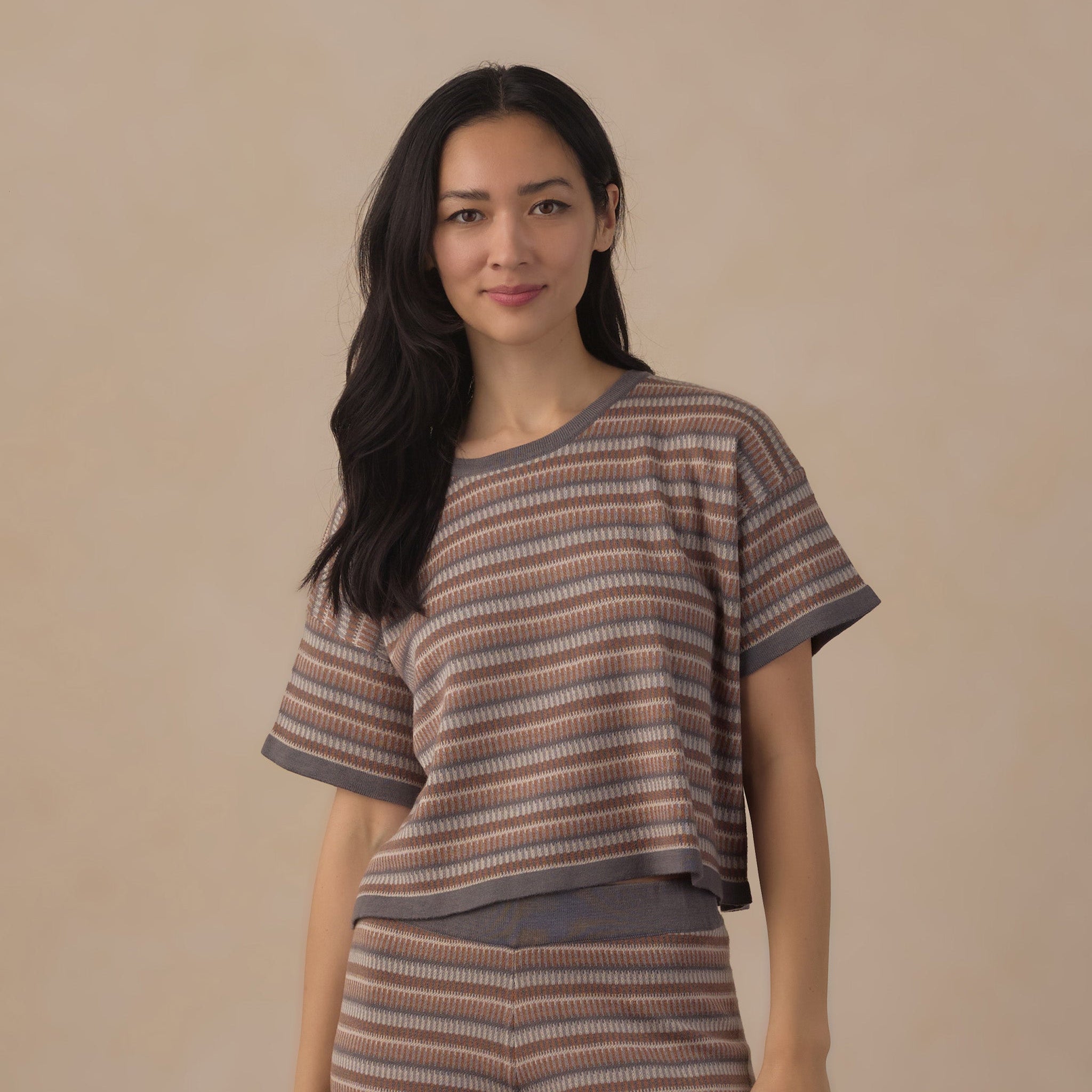 A woman modeling a matching two-piece knit loungewear set featuring a short-sleeve top and leggings with horizontal brown, beige, and gray stripes, set against a solid beige background.