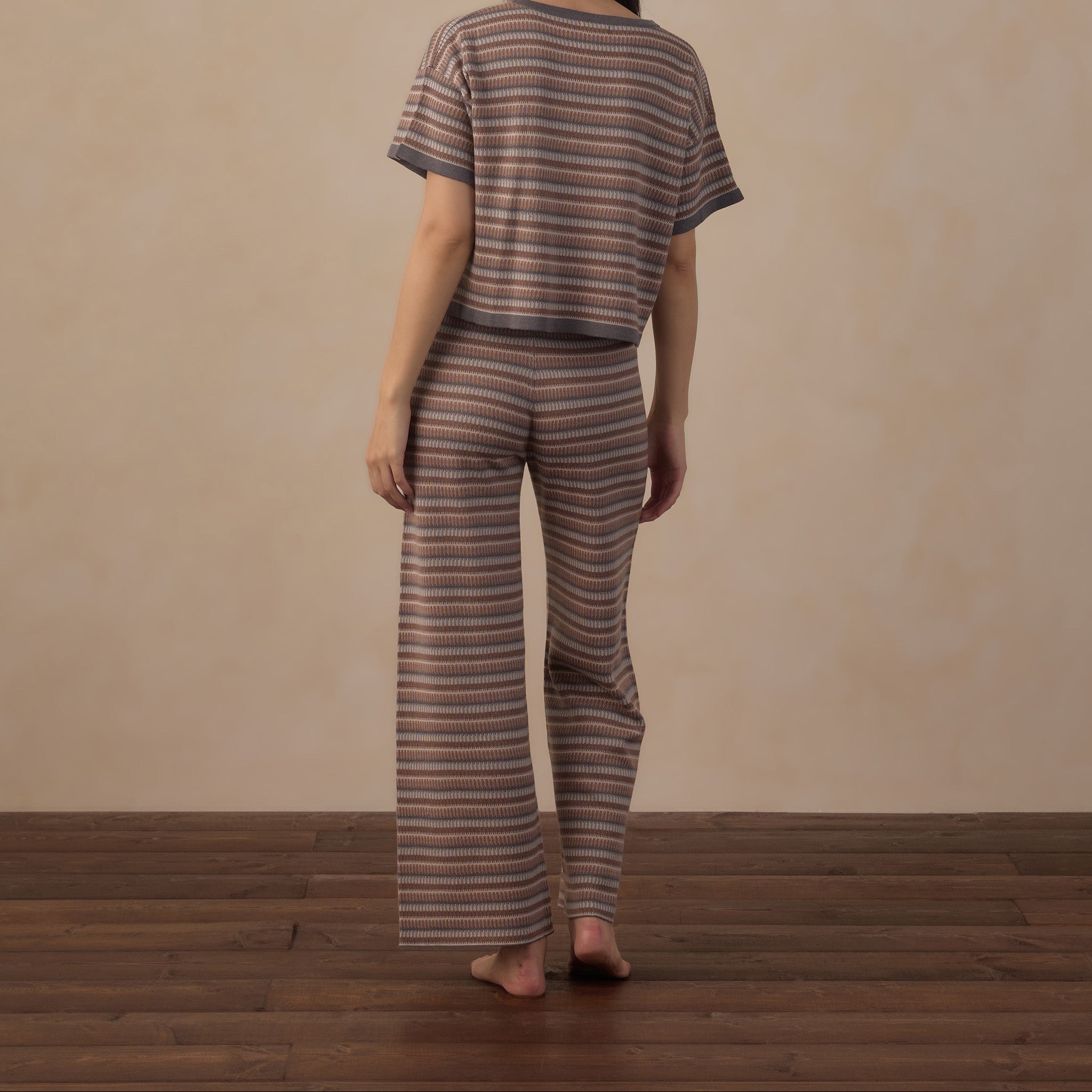 A woman stands with her back to the camera wearing a matching two-piece loungewear set with horizontal stripes in brown, beige, and muted red tones, set against a neutral beige wall and dark wood floor.