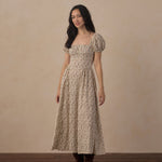 A woman stands in a studio wearing a long beige dress with a dense small floral print, puff sleeves, and a square neckline. She is also wearing dark brown boots against a warm beige backdrop and dark wood floor.