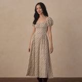 A woman stands in a studio wearing a long beige dress with a dense small floral print, puff sleeves, and a square neckline. She is also wearing dark brown boots against a warm beige backdrop and dark wood floor.