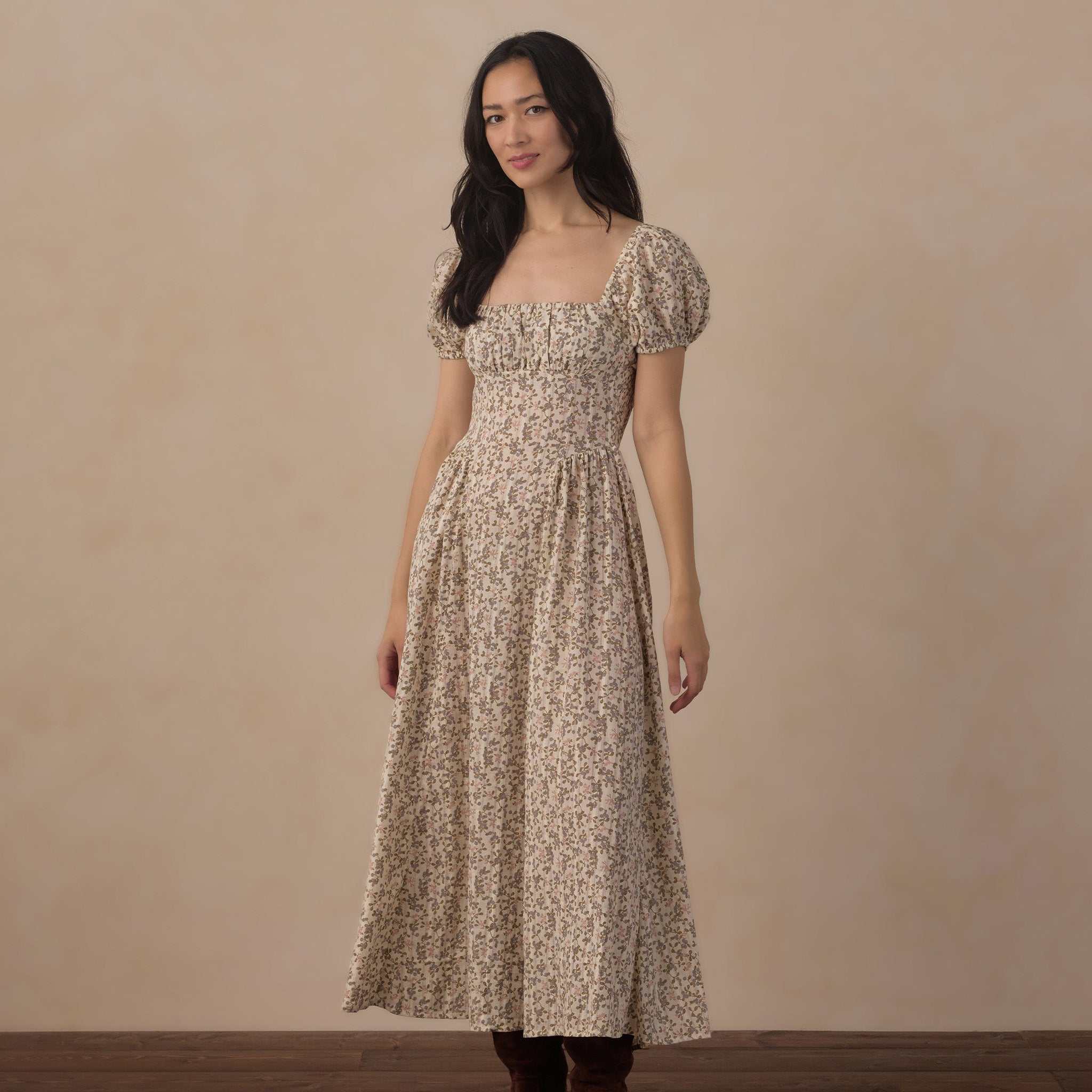 A woman stands in a studio wearing a long beige dress with a dense small floral print, puff sleeves, and a square neckline. She is also wearing dark brown boots against a warm beige backdrop and dark wood floor.