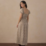 A woman modeling a beige and brown floral midi dress with puff sleeves and a smocked bodice, shown from the back against a neutral beige wall and wooden floor.