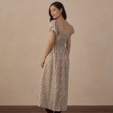 A woman modeling a beige and brown floral midi dress with puff sleeves and a smocked bodice, shown from the back against a neutral beige wall and wooden floor.
