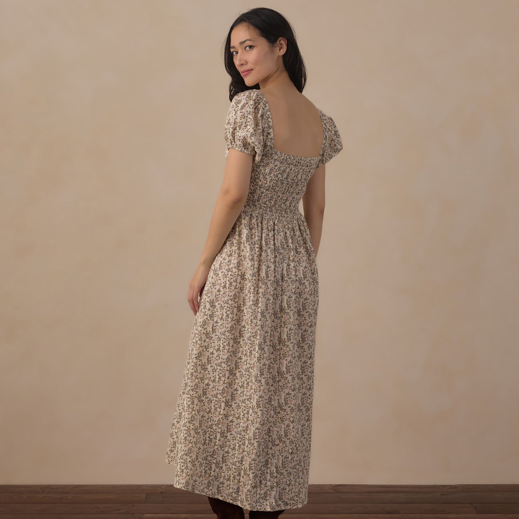 A woman modeling a beige and brown floral midi dress with puff sleeves and a smocked bodice, shown from the back against a neutral beige wall and wooden floor.