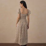 A woman stands with her back to the camera wearing a long-sleeved, smocked maxi dress with a beige and brown floral pattern, paired with brown boots, set against a neutral beige wall and dark wood floor.