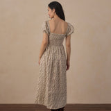 A woman stands with her back to the camera wearing a long-sleeved, smocked maxi dress with a beige and brown floral pattern, paired with brown boots, set against a neutral beige wall and dark wood floor.