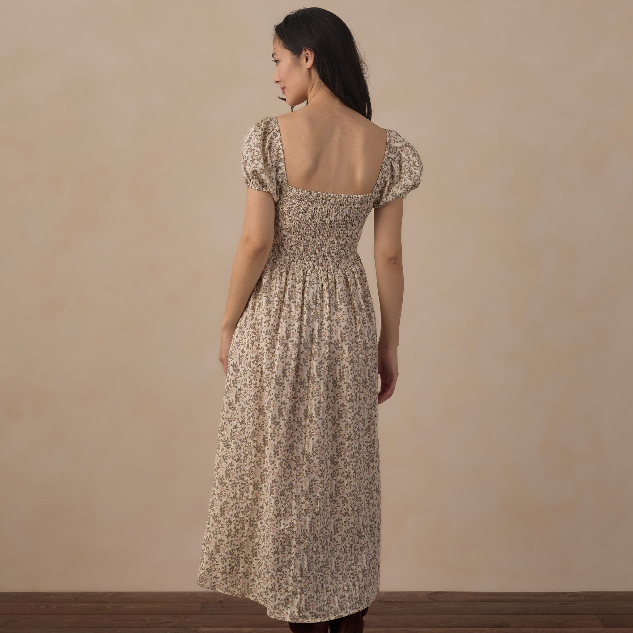 A woman stands with her back to the camera wearing a long-sleeved, smocked maxi dress with a beige and brown floral pattern, paired with brown boots, set against a neutral beige wall and dark wood floor.