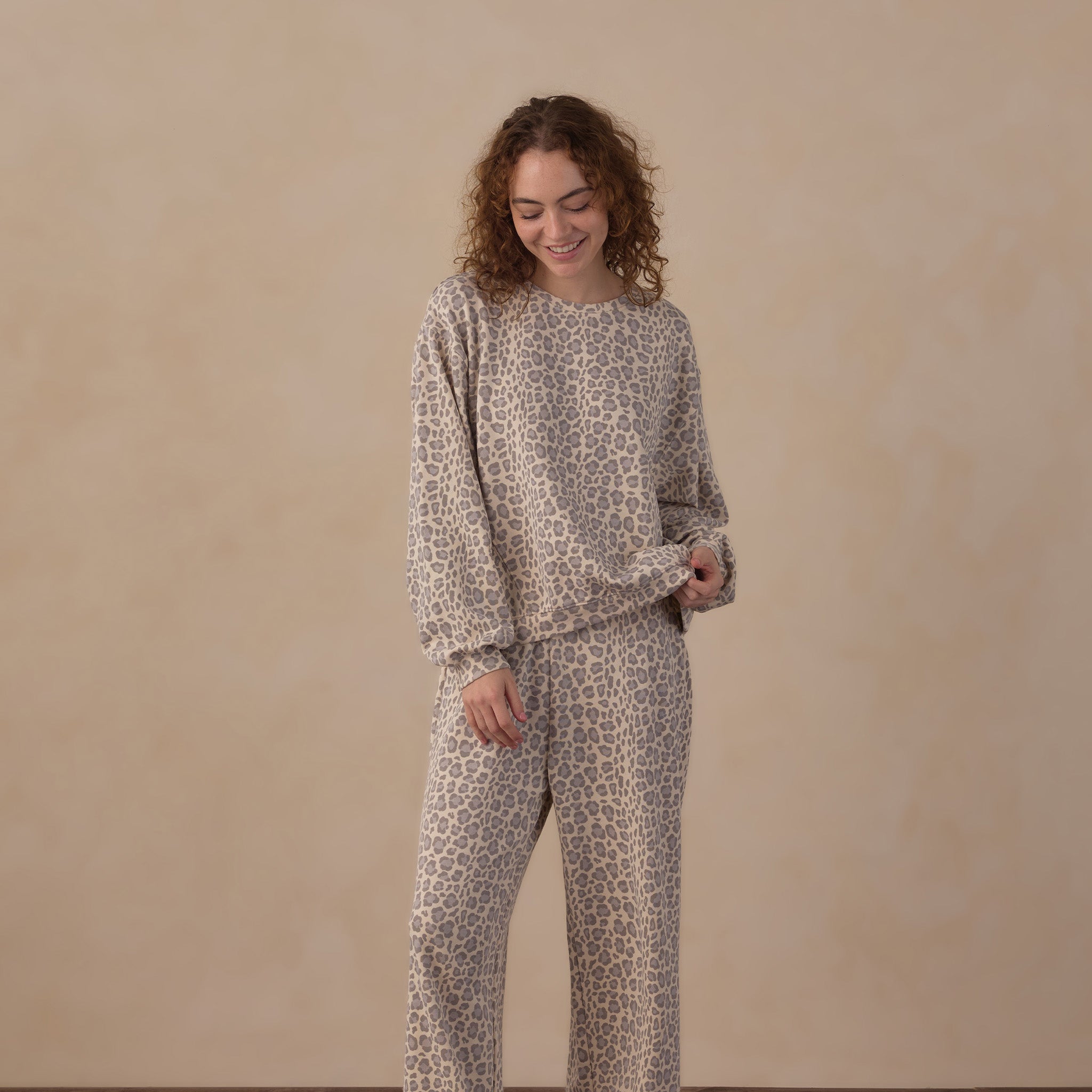A woman stands barefoot on a wooden floor against a beige wall, wearing a matching long-sleeve top and wide-leg pants set in a beige and brown animal print.