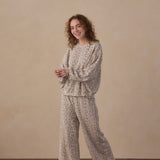A woman stands barefoot on a wooden floor against a beige wall, wearing a matching two-piece lounge set with a beige and brown abstract print. The set includes a long-sleeve top and wide-leg pants.