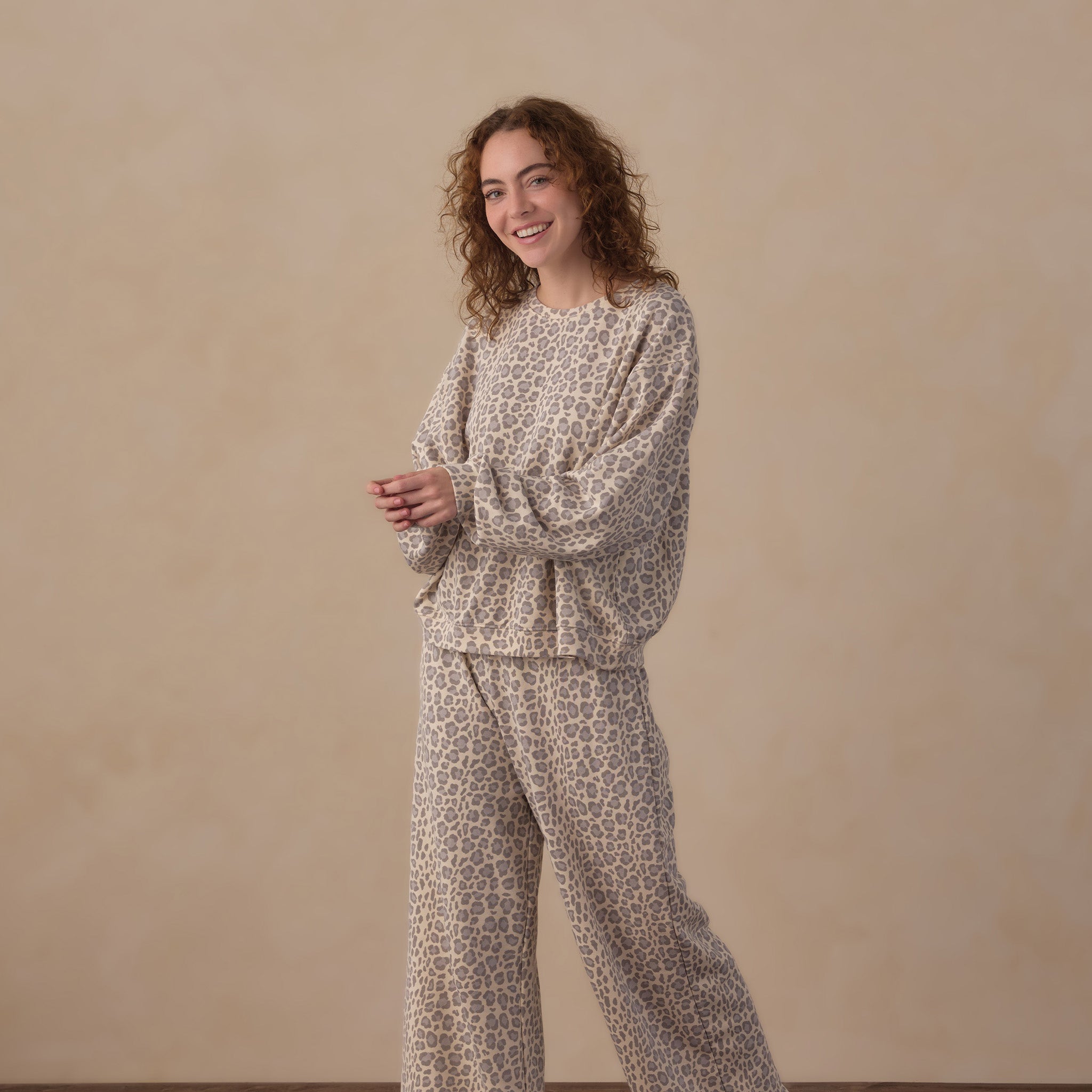 A woman stands barefoot on a wooden floor against a beige wall, wearing a matching two-piece lounge set with a beige and brown abstract print. The set includes a long-sleeve top and wide-leg pants.
