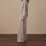 A woman with curly brown hair stands in profile wearing a long-sleeve maxi dress with a beige and brown abstract print, set against a neutral beige wall and dark wood floor.
