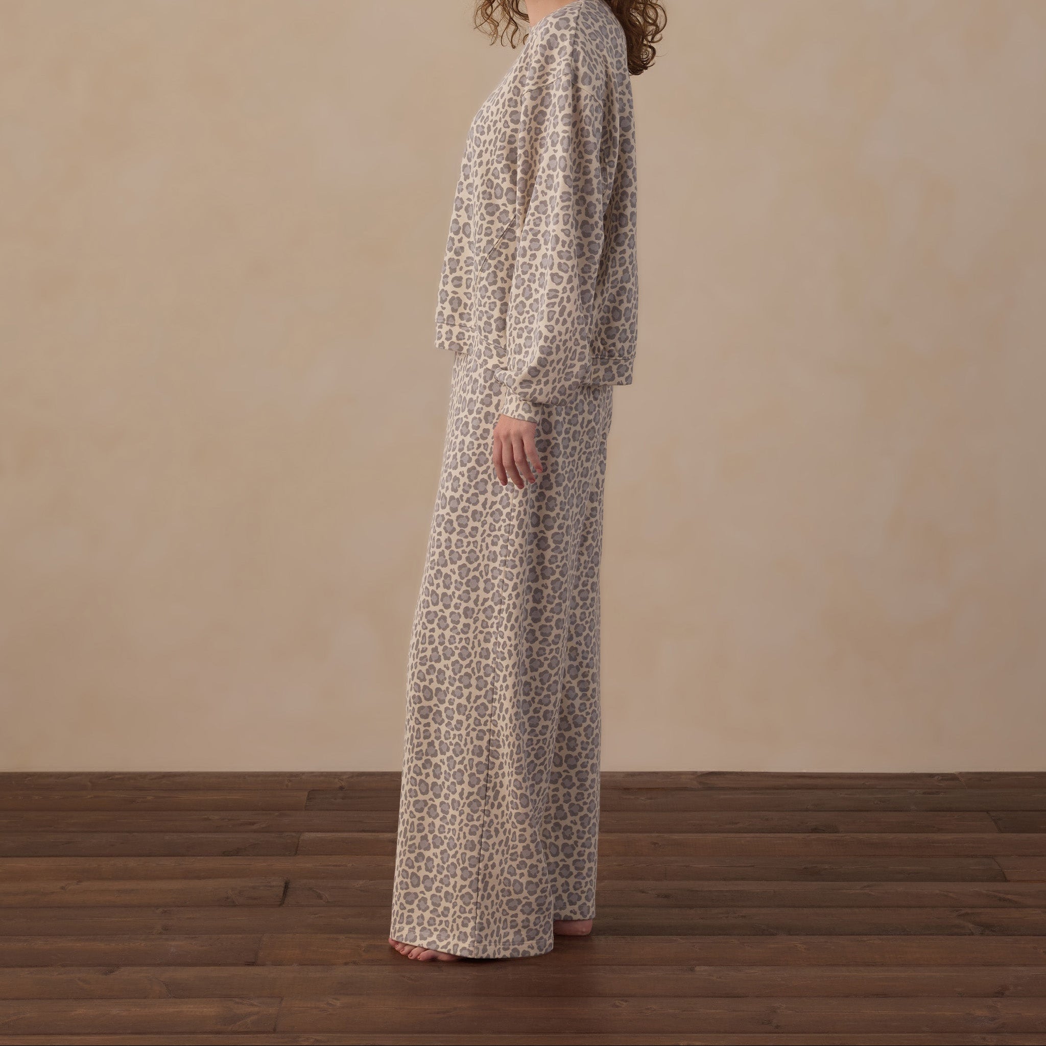 A woman with curly brown hair stands in profile wearing a long-sleeve maxi dress with a beige and brown abstract print, set against a neutral beige wall and dark wood floor.