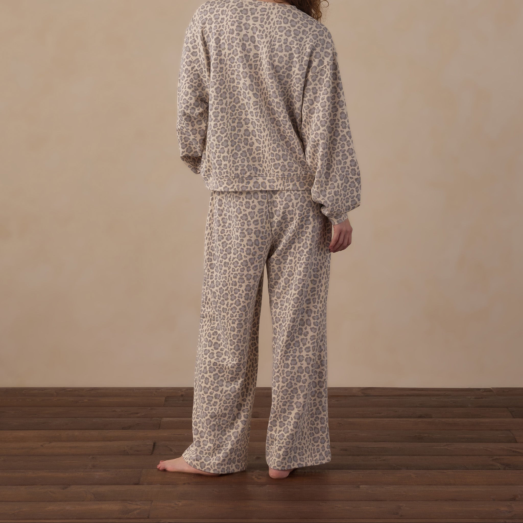 A person standing with their back to the camera wearing a matching two-piece pajama set with a beige and brown animal print pattern, set against a neutral beige wall and dark wood floor.