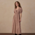 A woman stands in a studio wearing a dusty rose pink midi dress with a small floral print, short puff sleeves, and a gathered waist. She is also wearing dark brown suede ankle boots against a neutral beige textured backdrop and dark wood floor.