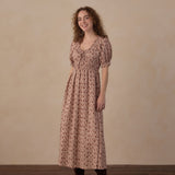 A woman stands in a studio wearing a dusty rose pink midi dress with a small floral print, short puff sleeves, and a gathered waist. She is also wearing dark brown suede ankle boots against a neutral beige textured backdrop and dark wood floor.