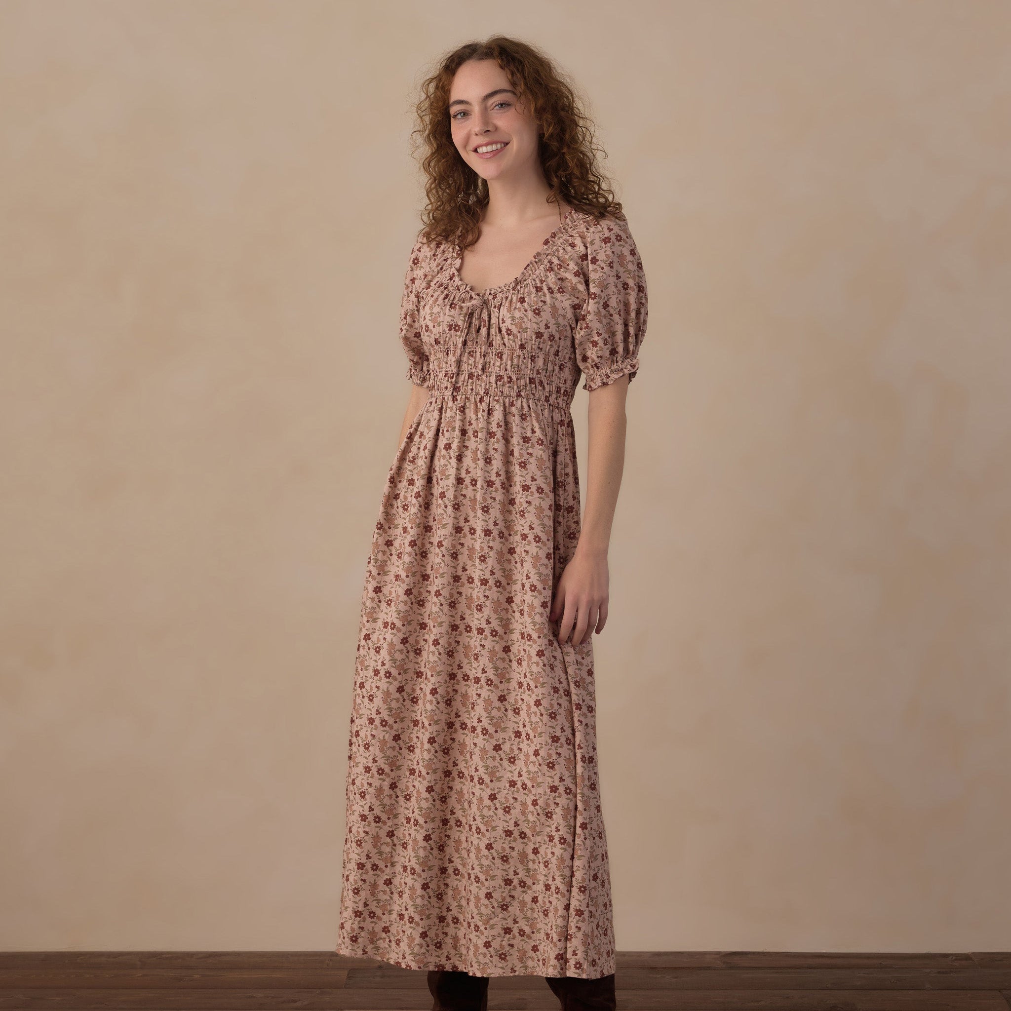 A woman stands in a studio wearing a dusty rose pink midi dress with a small floral print, short puff sleeves, and a gathered waist. She is also wearing dark brown suede ankle boots against a neutral beige textured backdrop and dark wood floor.