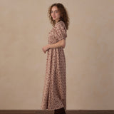 A woman with curly brown hair stands in profile wearing a knee-length puff-sleeve dress with a small floral print in muted pink and brown tones, paired with dark brown boots, against a warm beige studio wall and wooden floor.