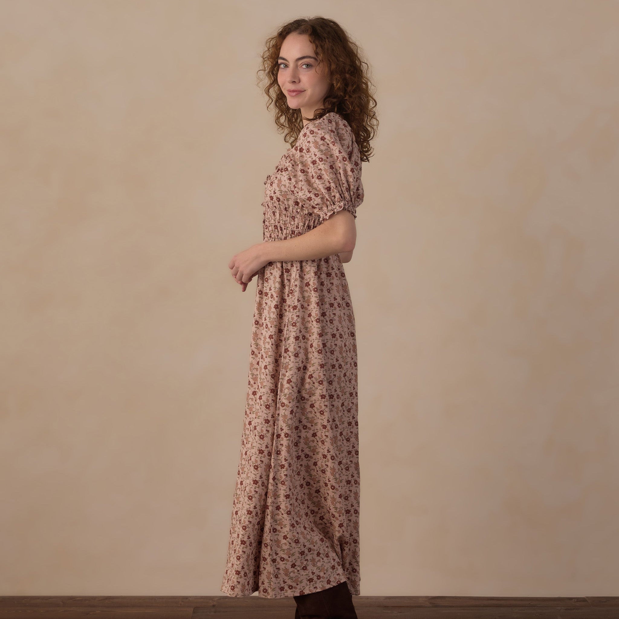 A woman with curly brown hair stands in profile wearing a knee-length puff-sleeve dress with a small floral print in muted pink and brown tones, paired with dark brown boots, against a warm beige studio wall and wooden floor.