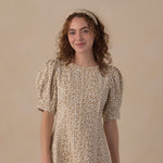 A woman with curly brown hair wearing a beige and brown leopard-print dress with puff sleeves and a matching headband, standing against a solid beige studio background.