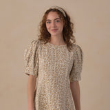 A woman with curly brown hair wearing a beige and brown leopard-print dress with puff sleeves and a matching headband, standing against a solid beige studio background.