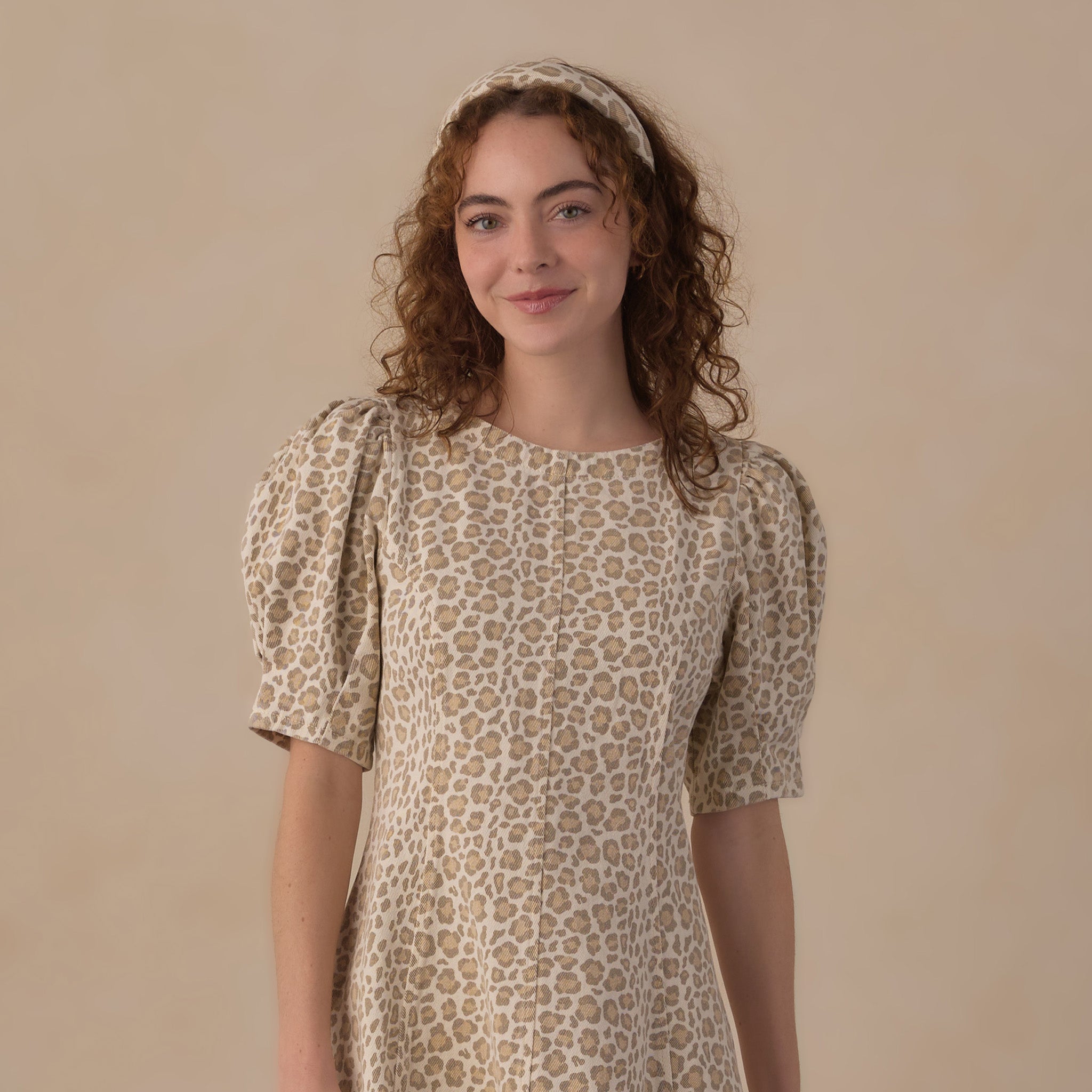 A woman with curly brown hair wearing a beige and brown leopard-print dress with puff sleeves and a matching headband, standing against a solid beige studio background.