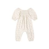A baby romper in a warm cream or off-white color with a delicate, small-scale floral print in muted blue and green. The romper features a smocked bodice, short puff sleeves, and leg openings that are gathered together at the ankles.
