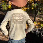 A child wearing a tan cowboy hat and a cream long-sleeve shirt with a distressed brown graphic on the back, standing outdoors in a wooded setting.