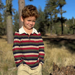 A young boy with curly brown hair stands outdoors in a wooded area wearing a long-sleeve polo shirt with horizontal stripes in red, navy, beige, and green, paired with khaki pants.