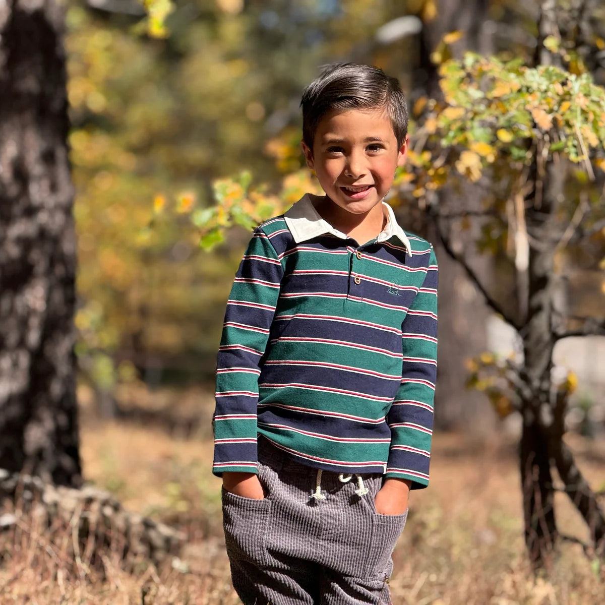 A young boy smiling outdoors in a wooded setting, wearing a long-sleeve collared polo shirt with horizontal stripes in green, navy, and white, paired with gray pants.
